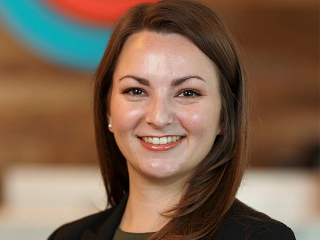 Professional headshot of smiling woman in black shirt against blurred indoor background.