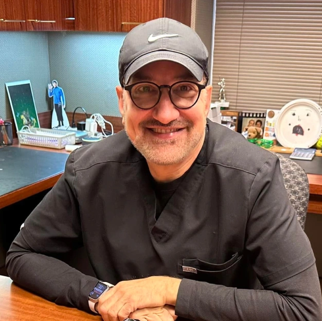 Man smiling at desk in orthodontics office wearing glasses and cap