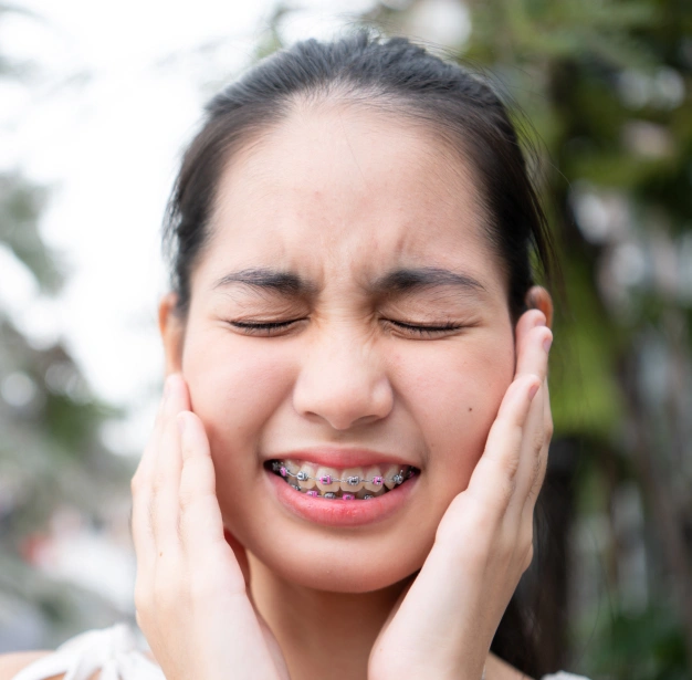 Young woman with braces holding her cheeks, smiling outdoors in natural setting.