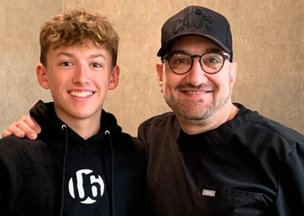 Two people smiling at camera in casual black clothing against beige background.
