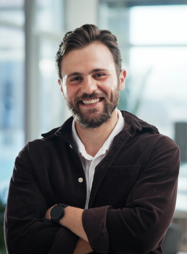 Professional headshot of smiling man in black jacket against modern office background