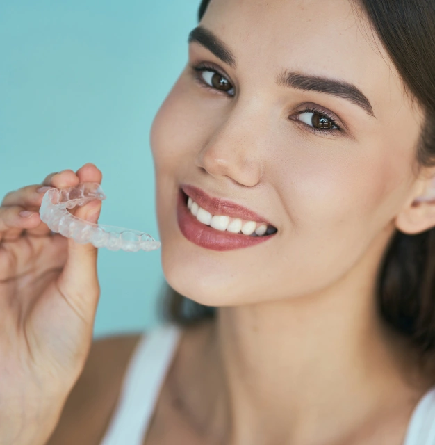 Woman smiling while holding clear aligner against blue background for orthodontics.