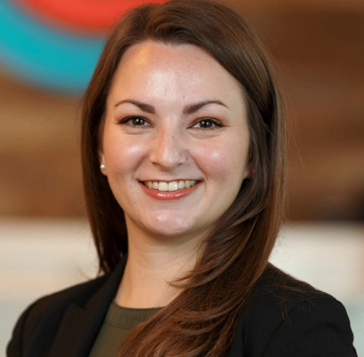 Professional headshot of smiling woman in black blazer against blurred office background.