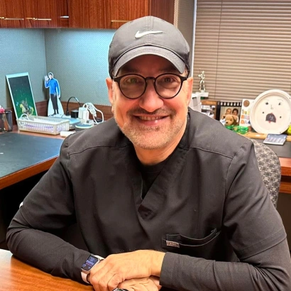 Man wearing glasses and cap smiling at desk in orthodontic office setting.