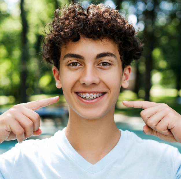 Young man smiling at camera, pointing to teeth, outdoor setting with green background.