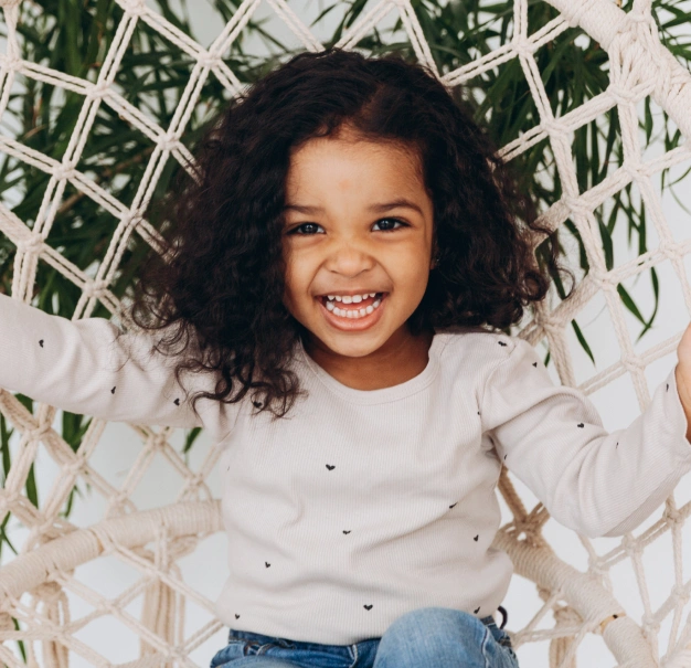 Happy child with curly dark hair smiling at camera in white chair with green foliage backdrop.