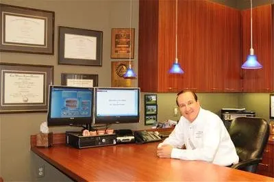 Orthodontist sits at desk in modern office with computers and diplomas on wall.