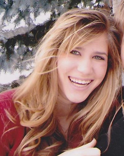 Smiling woman with reddish-blonde hair against snowy evergreen background, wearing burgundy top.