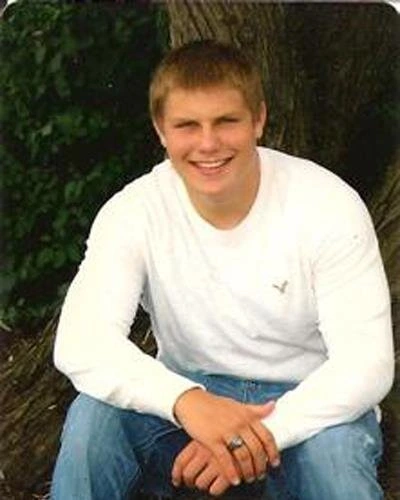 Young man in white long-sleeve shirt and jeans smiling at camera against green foliage background.
