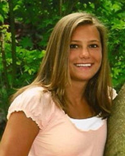 Woman smiling at camera wearing pink shirt, green foliage background, professional portrait setting.