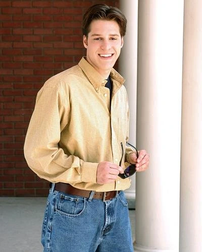 Young man in yellow shirt holding clipboard smiling against brick wall and white column.